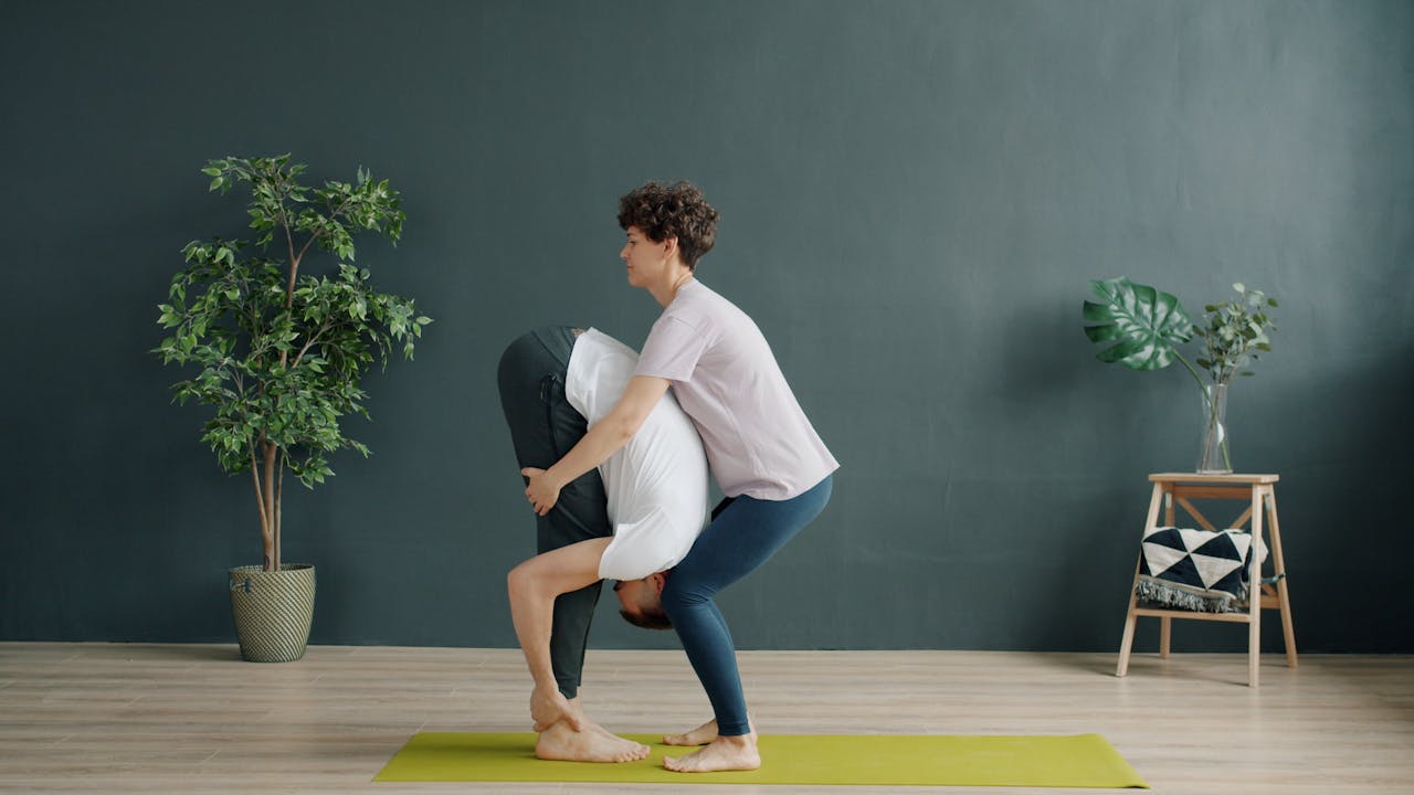 About Yoga instructor helps student achieve the perfect forward bend pose indoors.